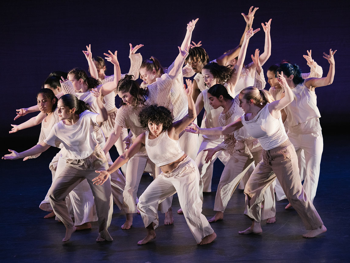 A group of college students dressed in light-colored clothes performs a choreographed dance on a dark stage